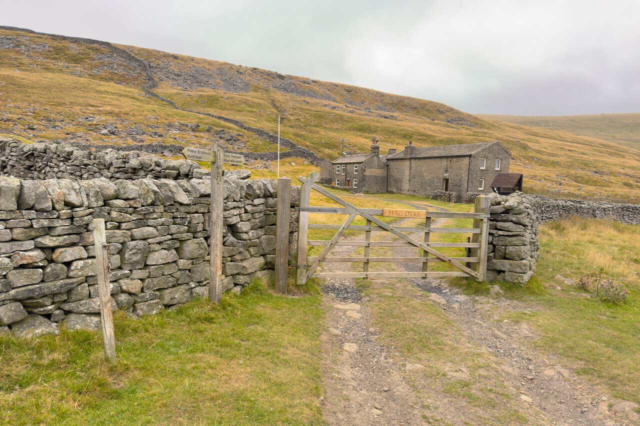 A wooden gate marked Hag Dyke, with a footpath signpost and the hostel buildings on the fellside beyond. A highlight on this Great Whernside circular walk.