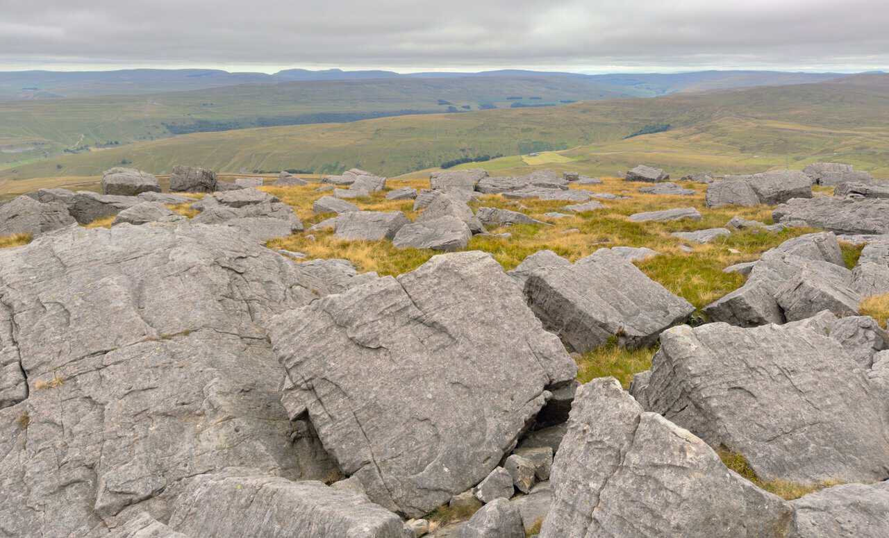 Large gritstone boulders on the summit plateau, with distant fells along the western skyline.