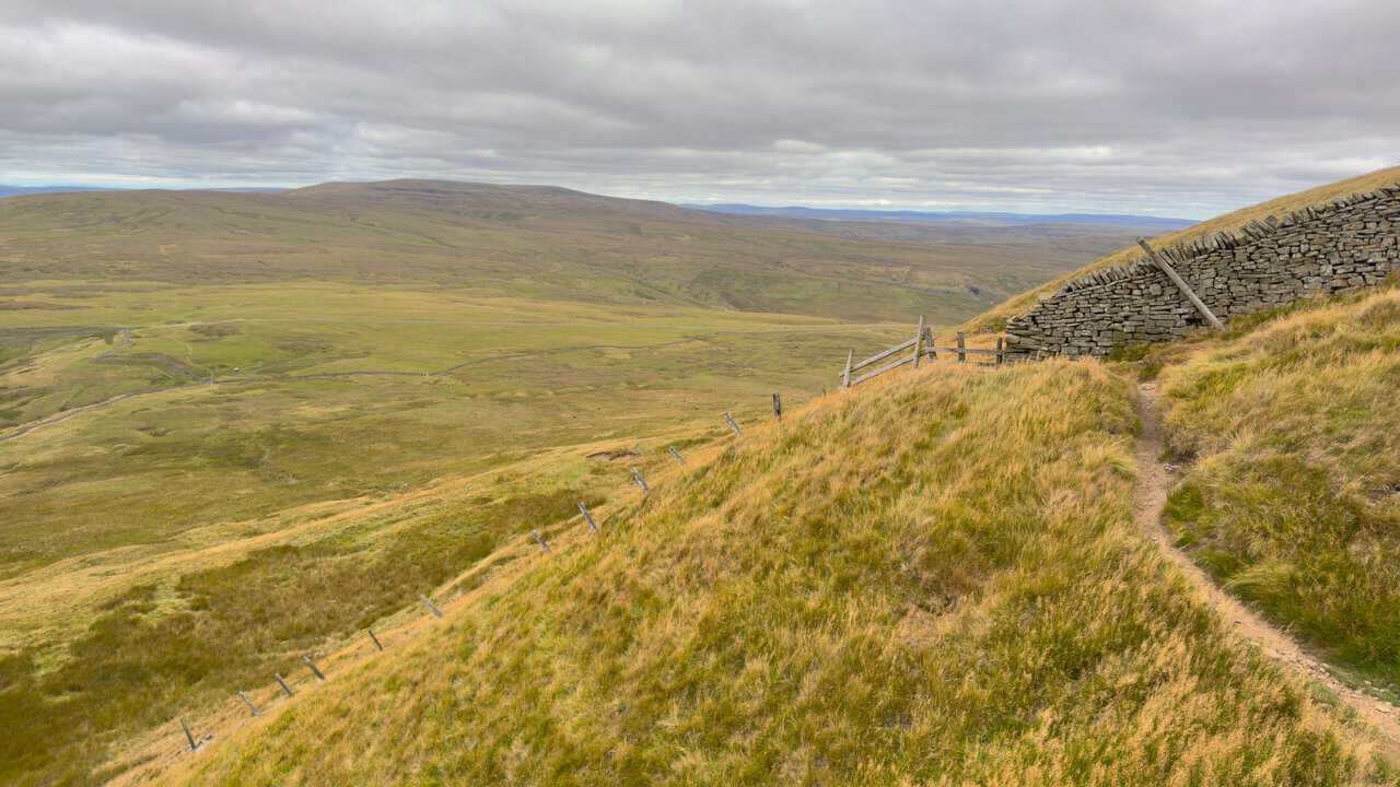 The footpath dropping off the mountainside beside a fence, with Buckden Pike in the distance.