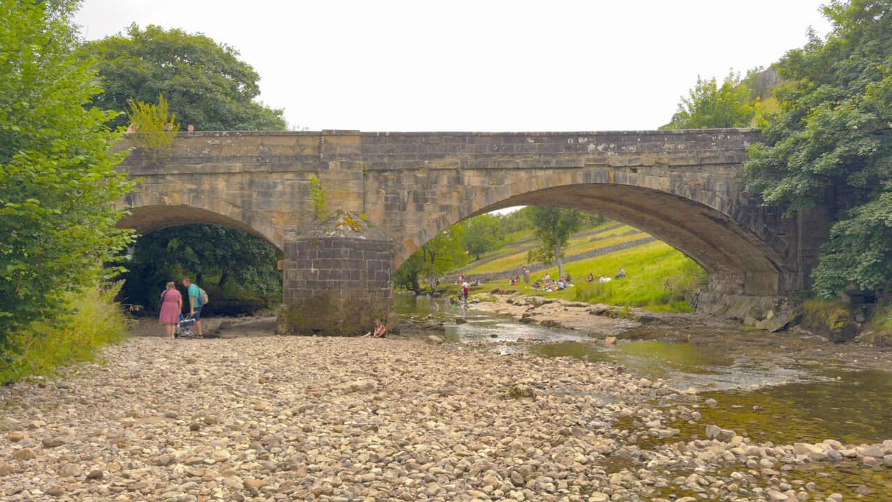 The stone road bridge carrying the B6160 over the River Wharfe, with families on the shingle below.