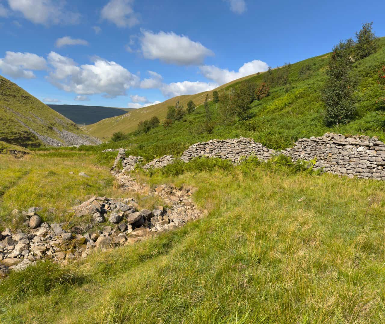 The tumbled remains of a stone wall beside the path, with a broad scree slope spilling down the far side of the valley.