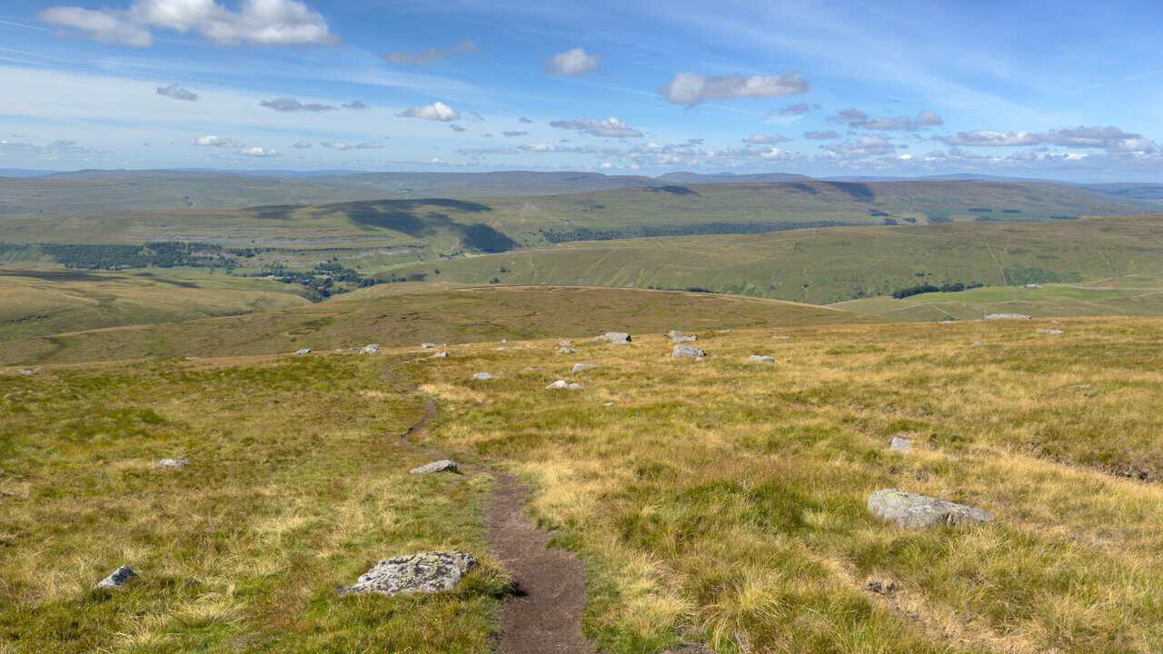 A thin path winding across grassy moorland scattered with boulders, with the dales stretching away behind.