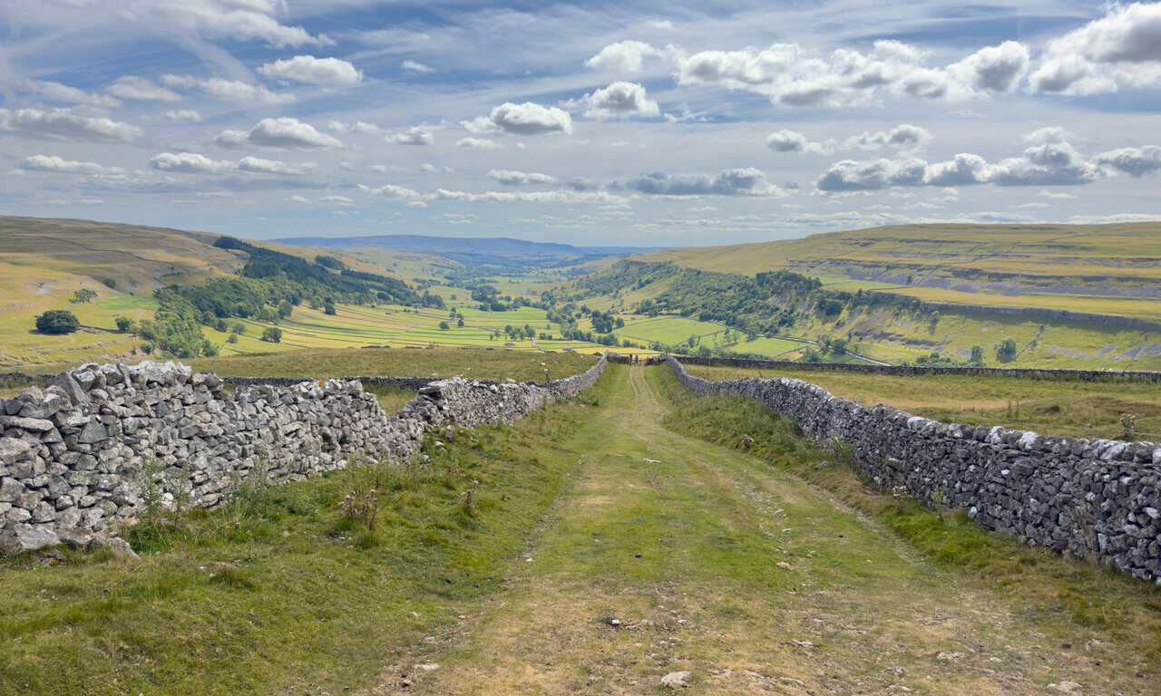 The walled green track descending into a widening Wharfedale, with pastures and limestone scars ahead.