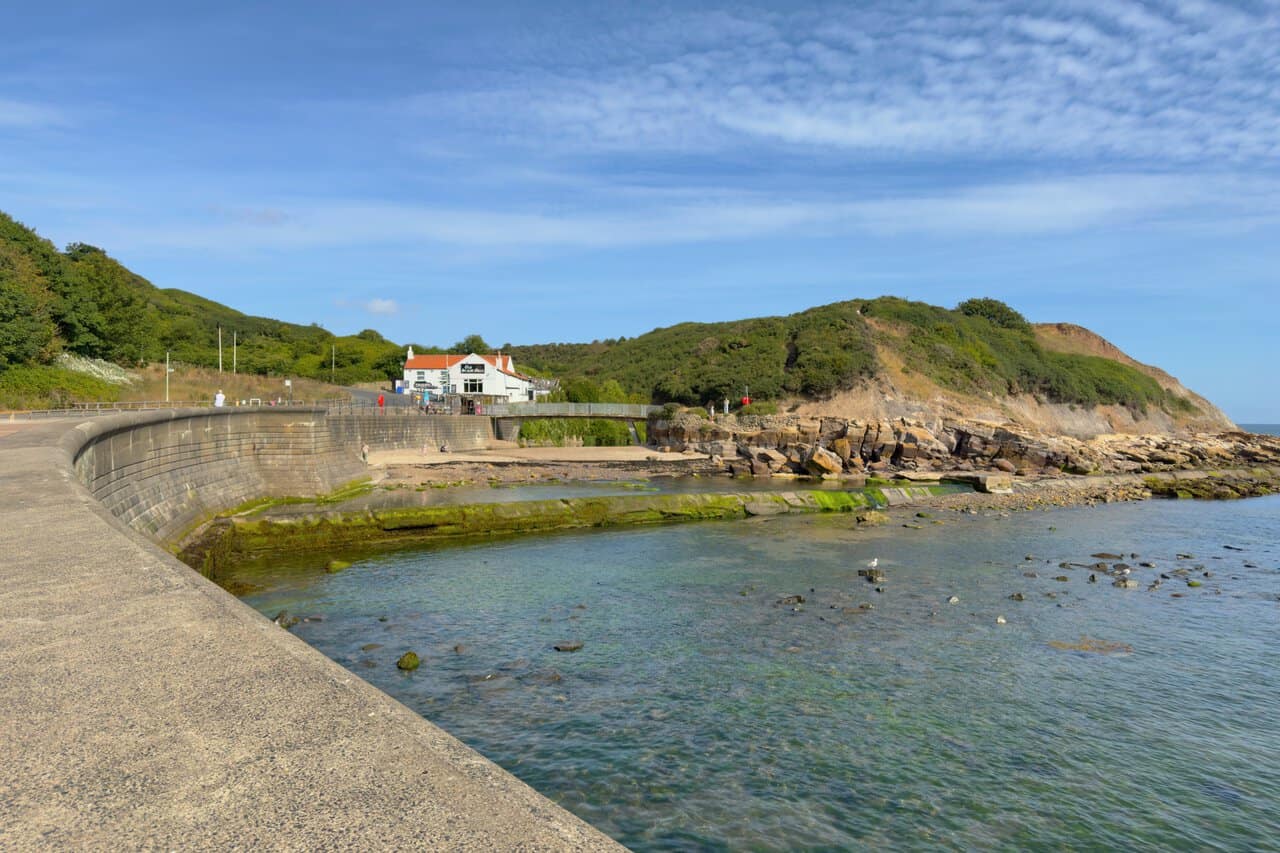 The Old Scalby Mills, a whitewashed pub with an orange pantile roof, stands at the northern end of North Bay, with a footbridge and rocky shore in front of it.