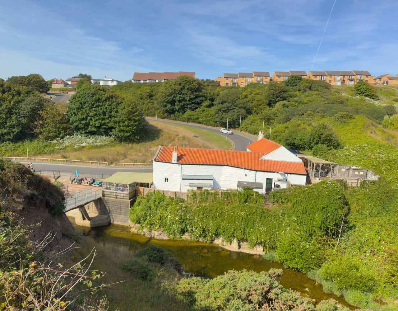 Looking down on Scalby Mills from the cliff steps, showing the old mill building, the footbridge over the beck and the coast road curling above.