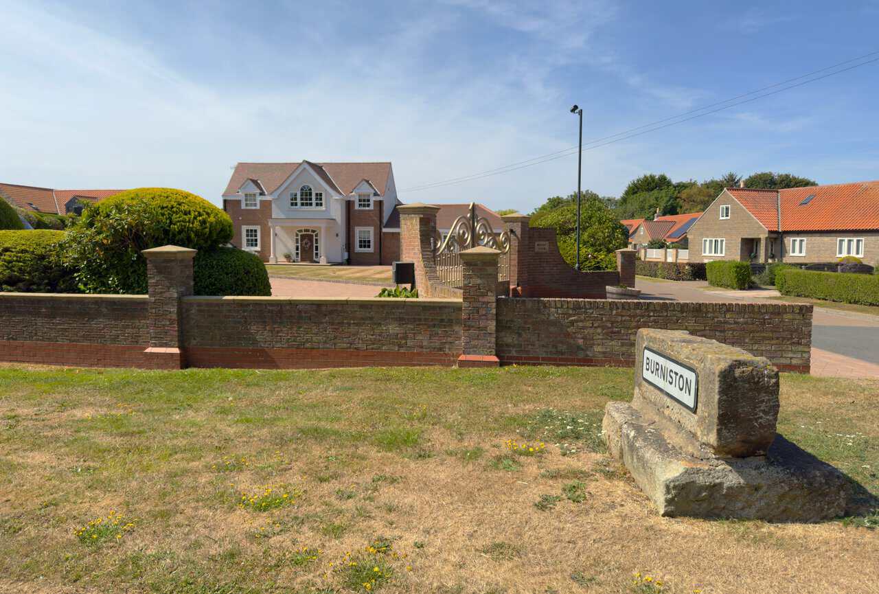 The Burniston village sign set on a weathered block of stone at the roadside, with the house called Mount View behind its brick wall.