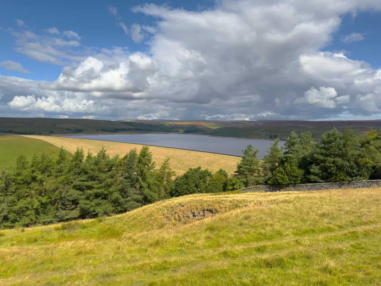 Grimwith Reservoir seen from a grassy hillside, with sunlight on the embankment and dark moorland beyond.