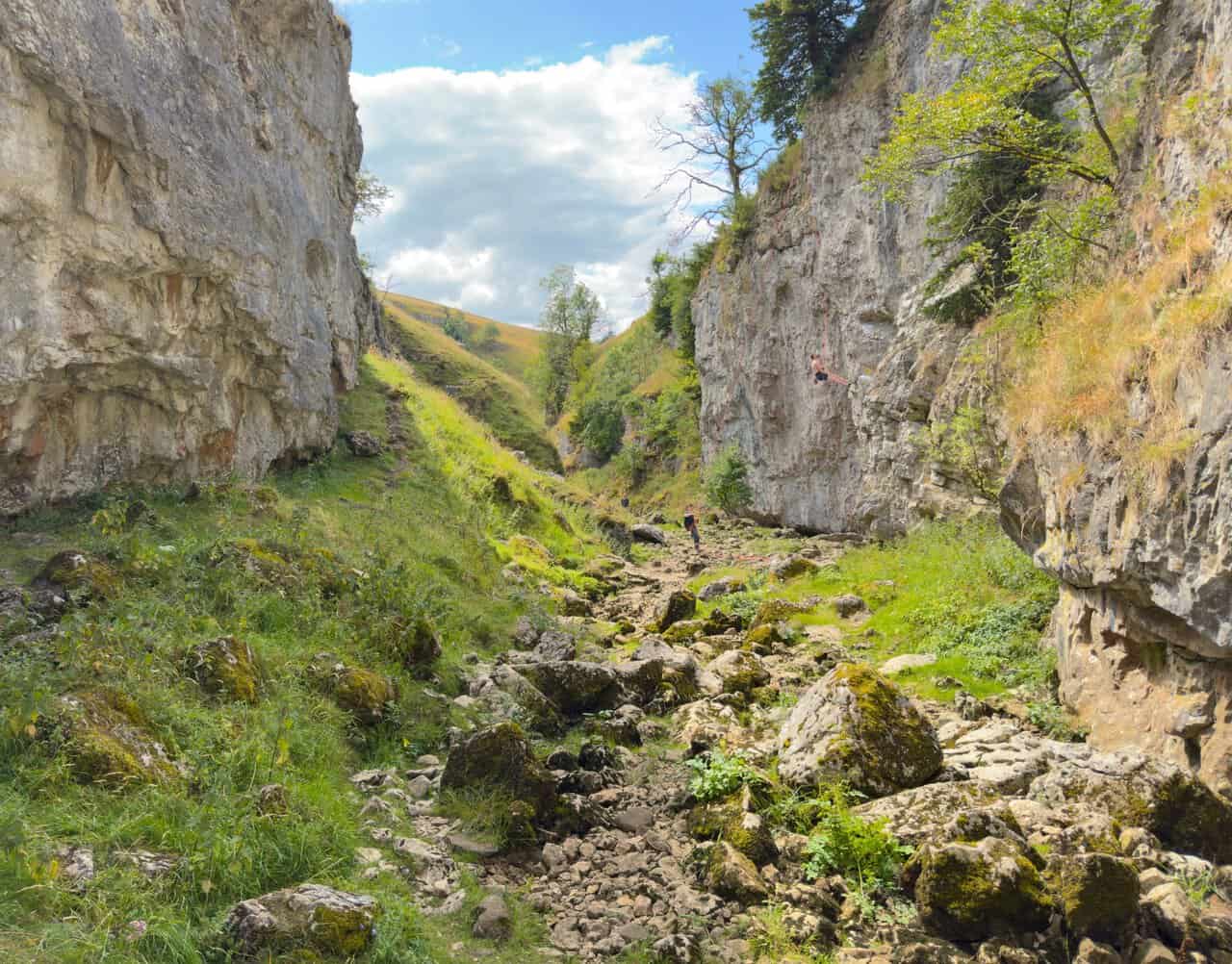 Two rock climbers on the limestone wall in the heart of the Trollers Gill walk ravine, one climbing and one holding the ropes.