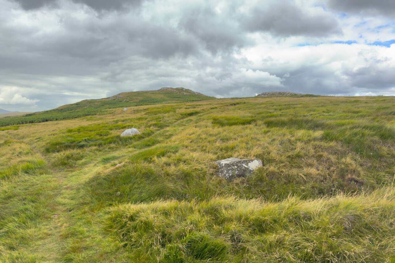 A faint grassy path climbing through moor grass towards the rocky outcrops of Nursery Knot.