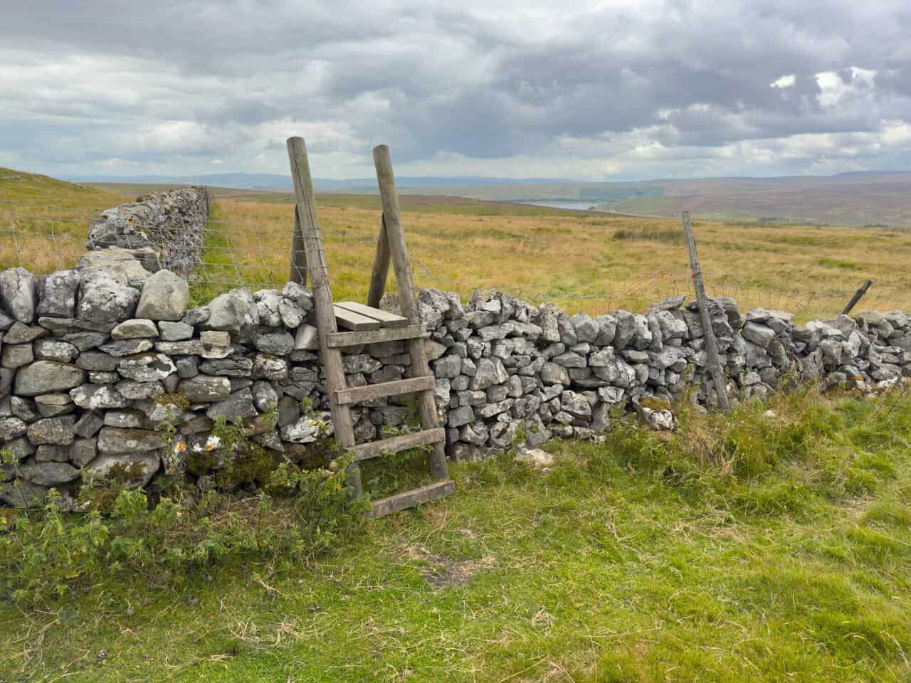 A wooden stile over a dry stone wall, with Grimwith Reservoir appearing in the distance beyond.