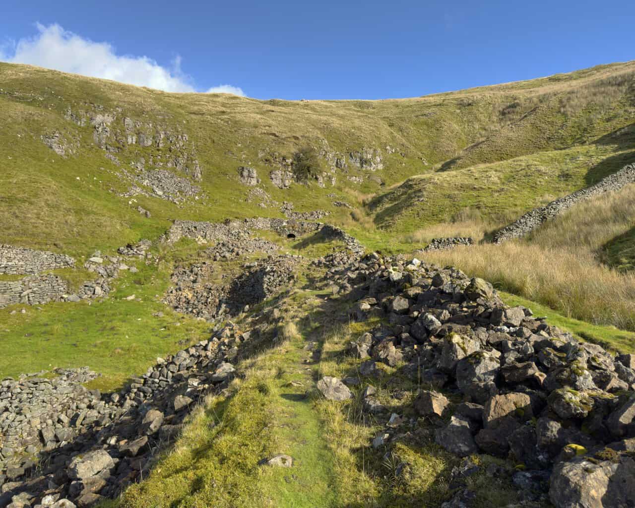 A grassy path leading through ruined walls and rock debris at the old Buckden Lead Mine workings.