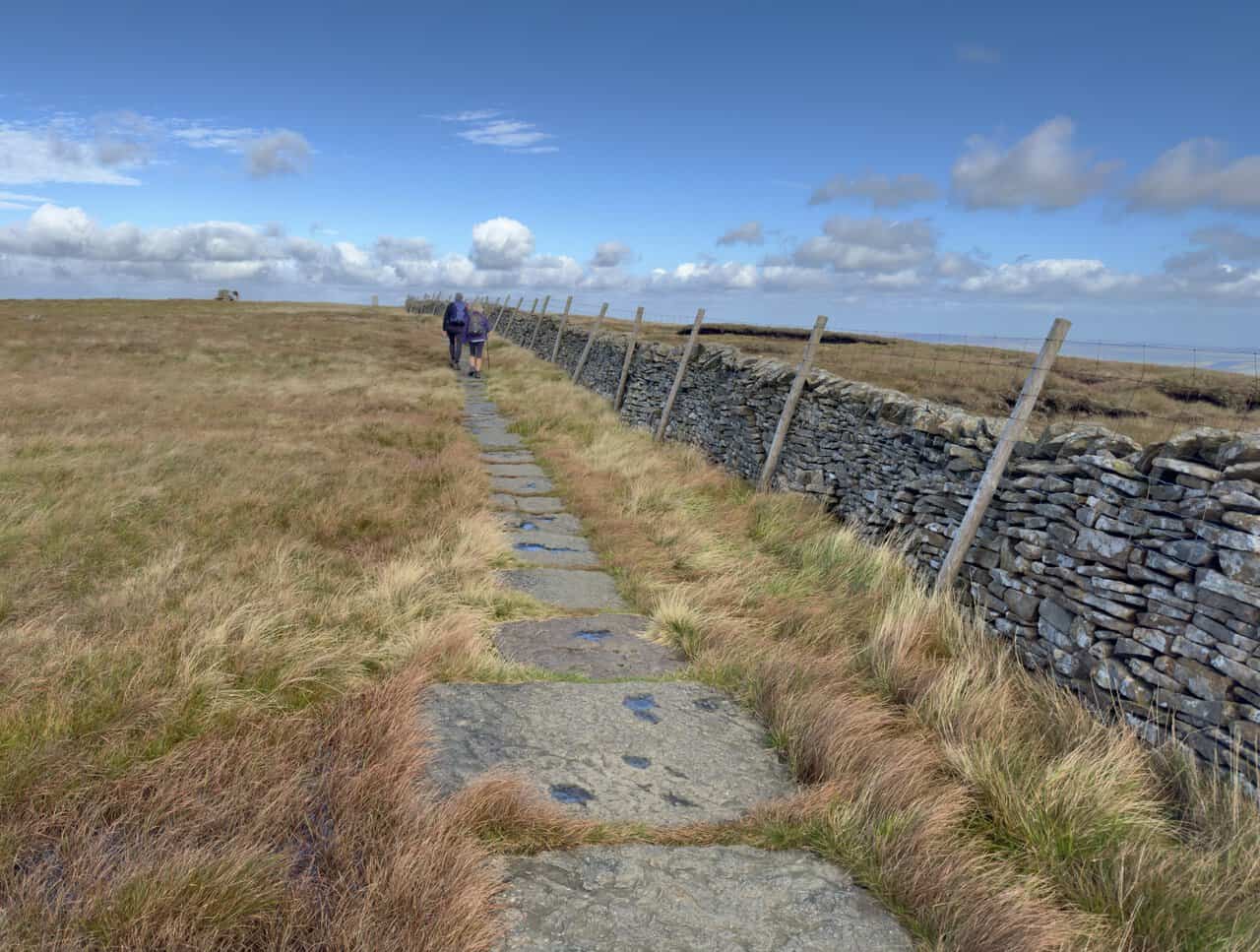 Two walkers on the flagged path beside a dry-stone wall near the summit of Buckden Pike.