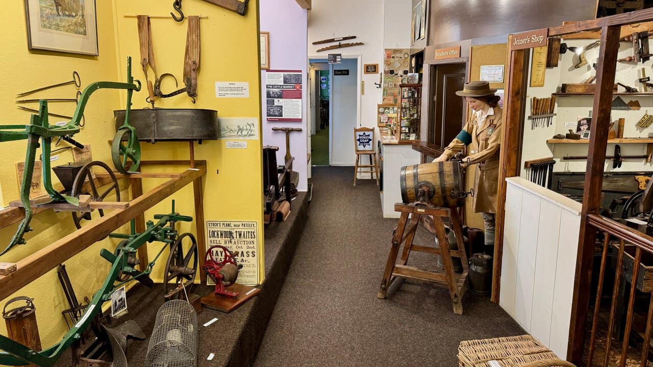 The Agriculture Room at Nidderdale Museum, with green farm implements, a Land Girl figure at a butter churn and shop fronts along the corridor.