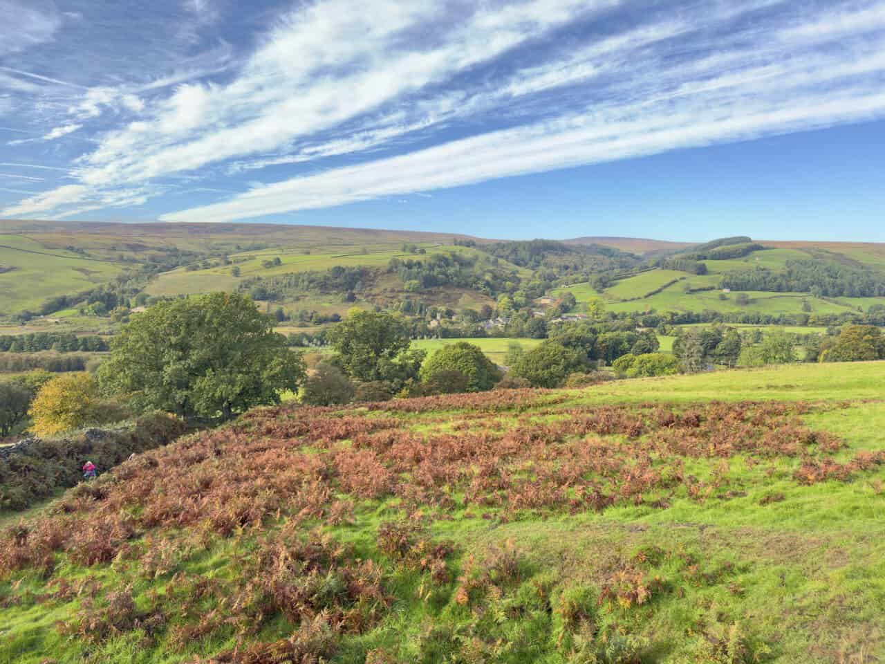 Looking back over russet bracken and green fields towards the village at the start of the Ramsgill walk, with wooded slopes and moorland beyond.