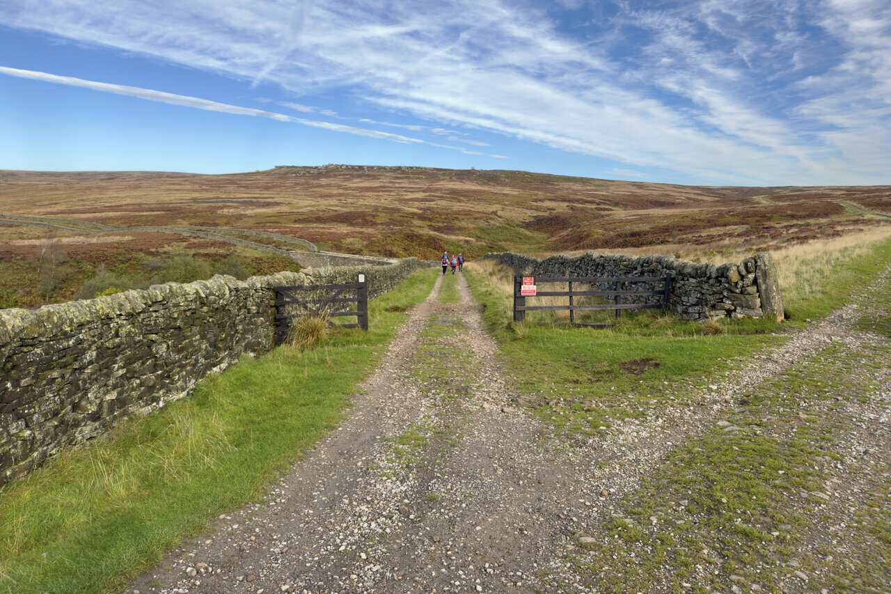 A moorland track junction with wooden gates in the dry-stone walls, and walkers heading towards Sypeland Crags on the skyline.