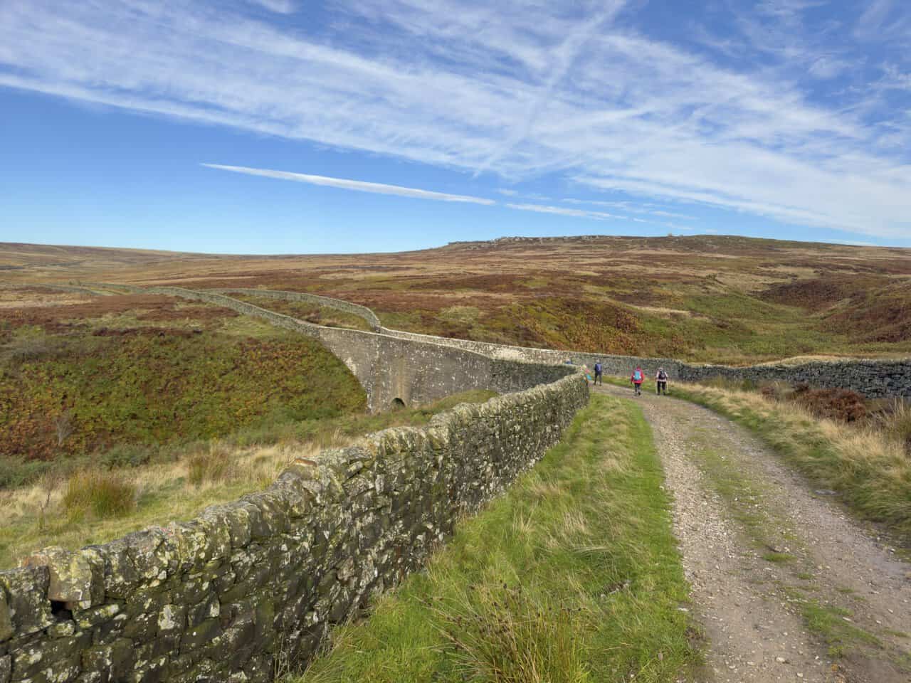A curving dry-stone wall crossing a stone bridge over Lul Beck, with walkers on the Ramsgill walk following the gravel track beside it.