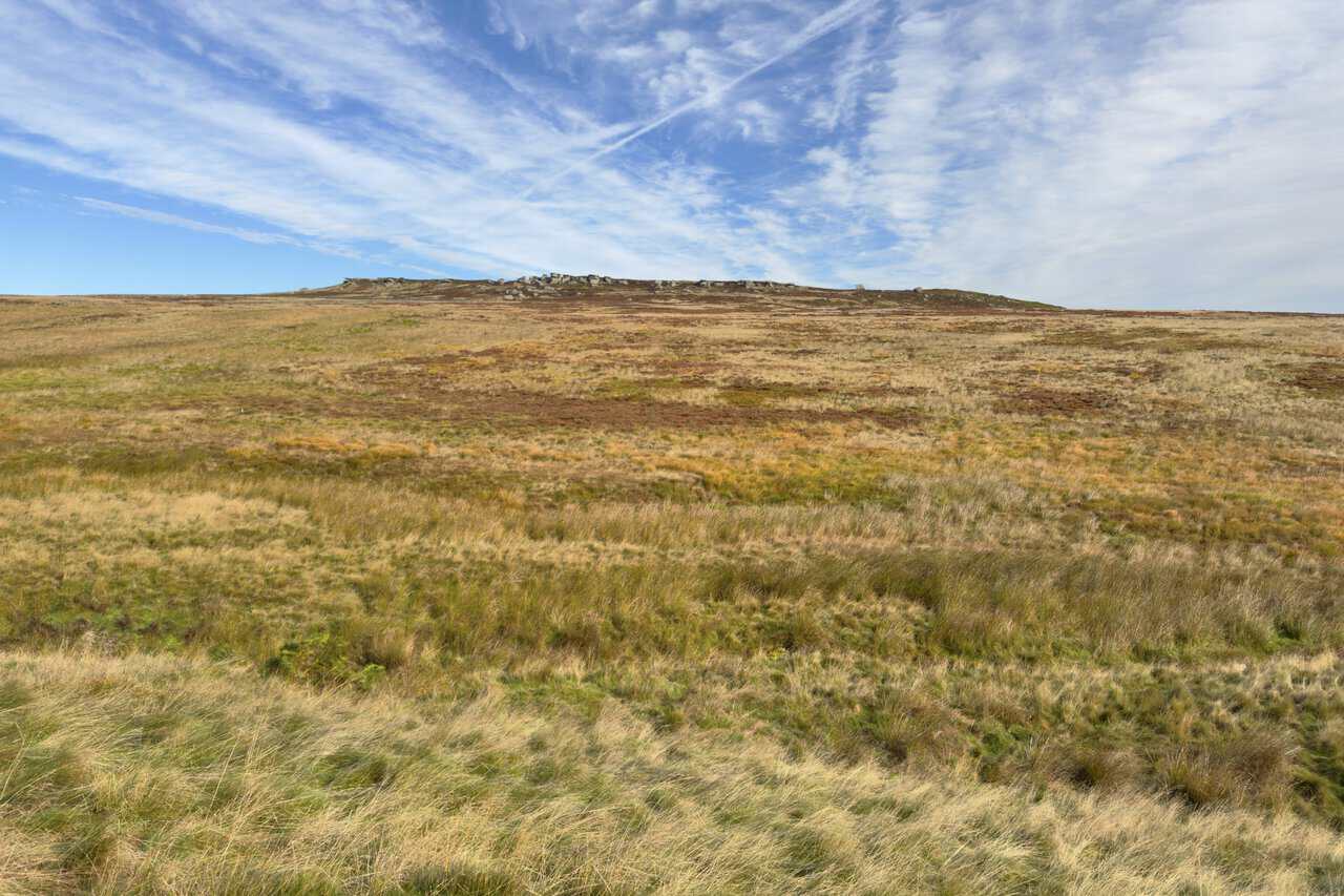 Sypeland Crags on the skyline above a broad expanse of moor grass, rushes and heather under a streaky blue sky.
