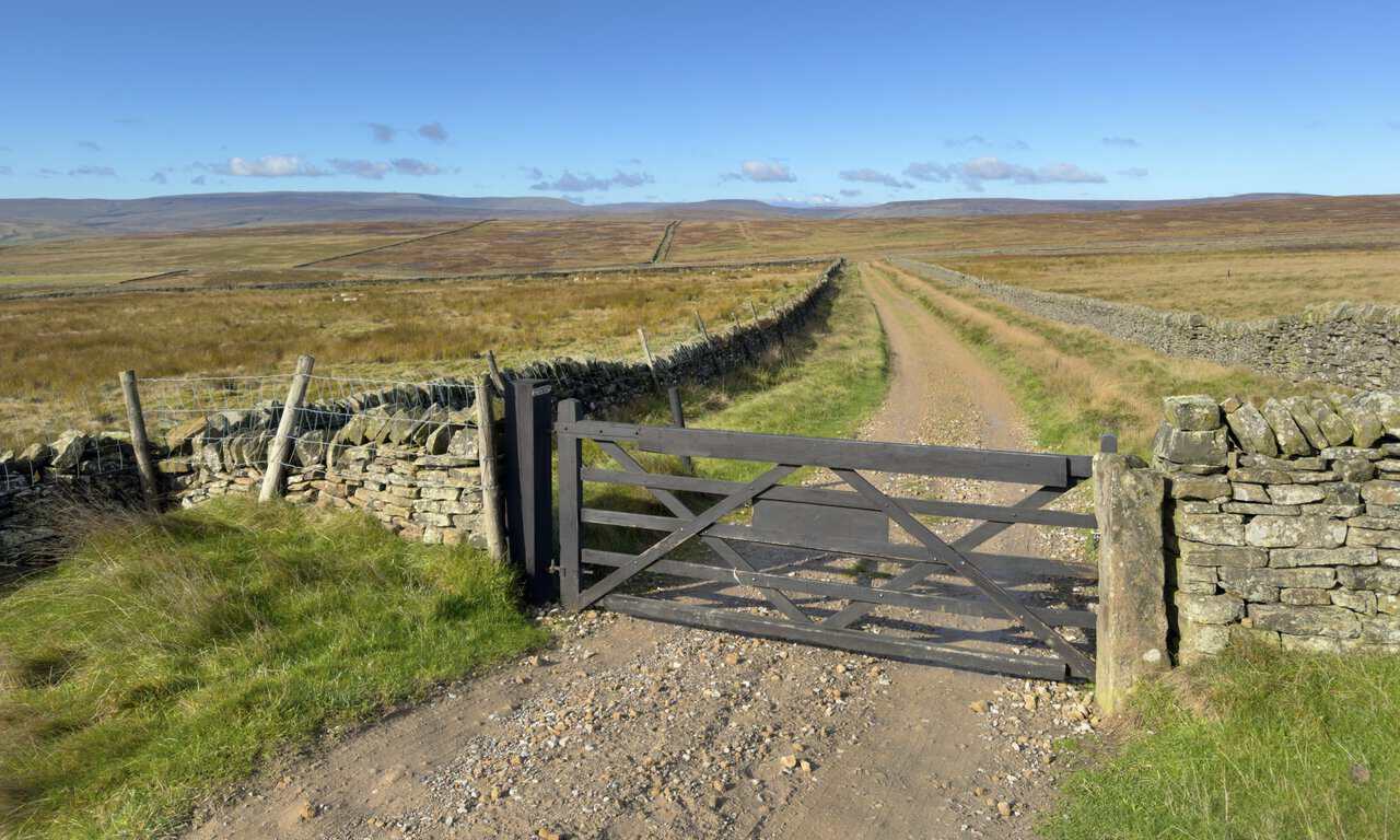 A dark wooden gate across a gravel track between dry-stone walls, with moorland and distant hills beyond.