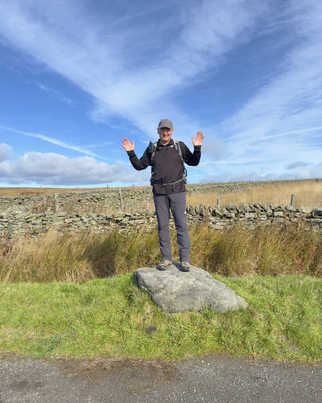 A smiling walker in a cap and rucksack standing on a roadside boulder during a coffee stop on the Ramsgill walk, with moorland behind.