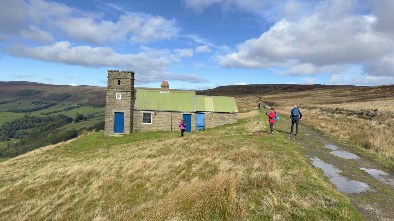 The Thrope Edge Shooting House, a stone building with a castellated tower, blue doors and a green roof, beside a moorland track with walkers passing.