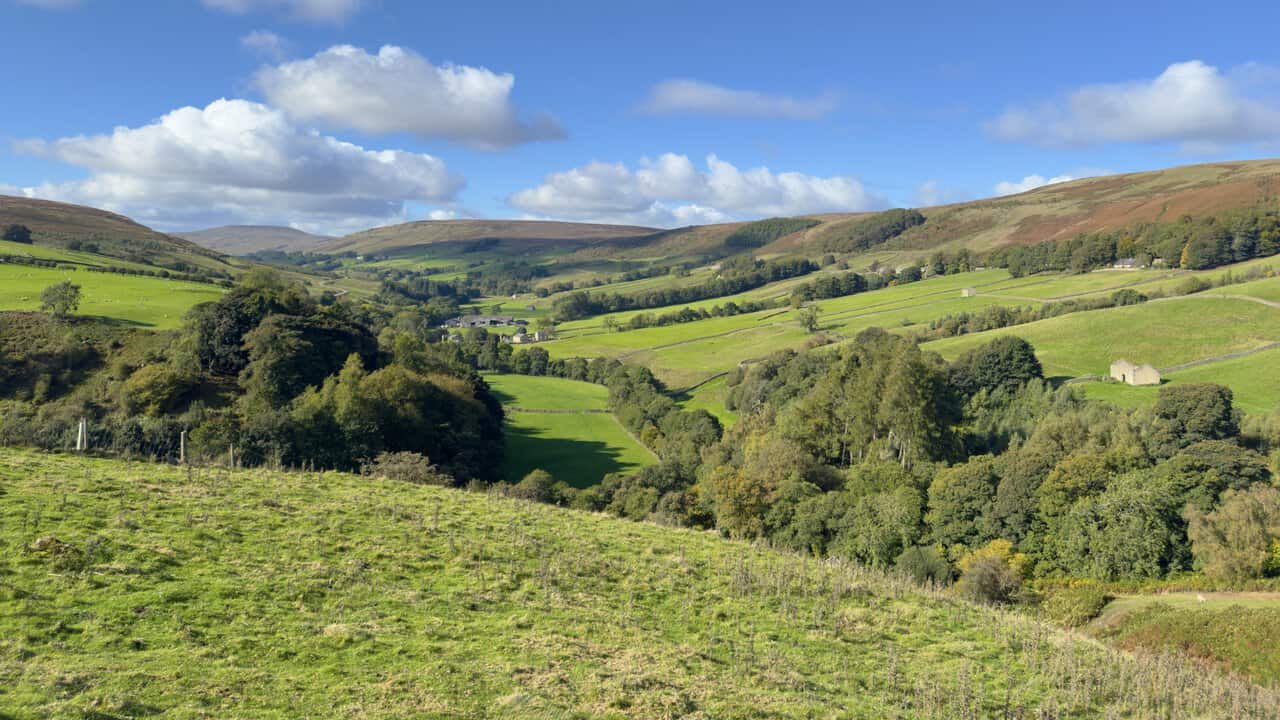 Green fields, dry-stone walls, field barns and woodland along the valley floor, beneath russet moorland slopes and white clouds.