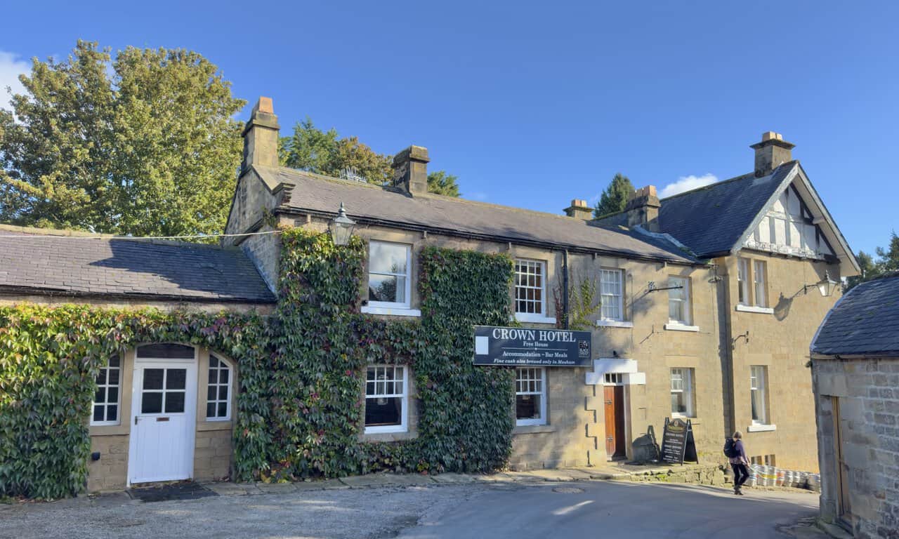 The stone-built Crown Hotel in Lofthouse, partly covered in creeper, with its sign above the windows and a walker passing by.