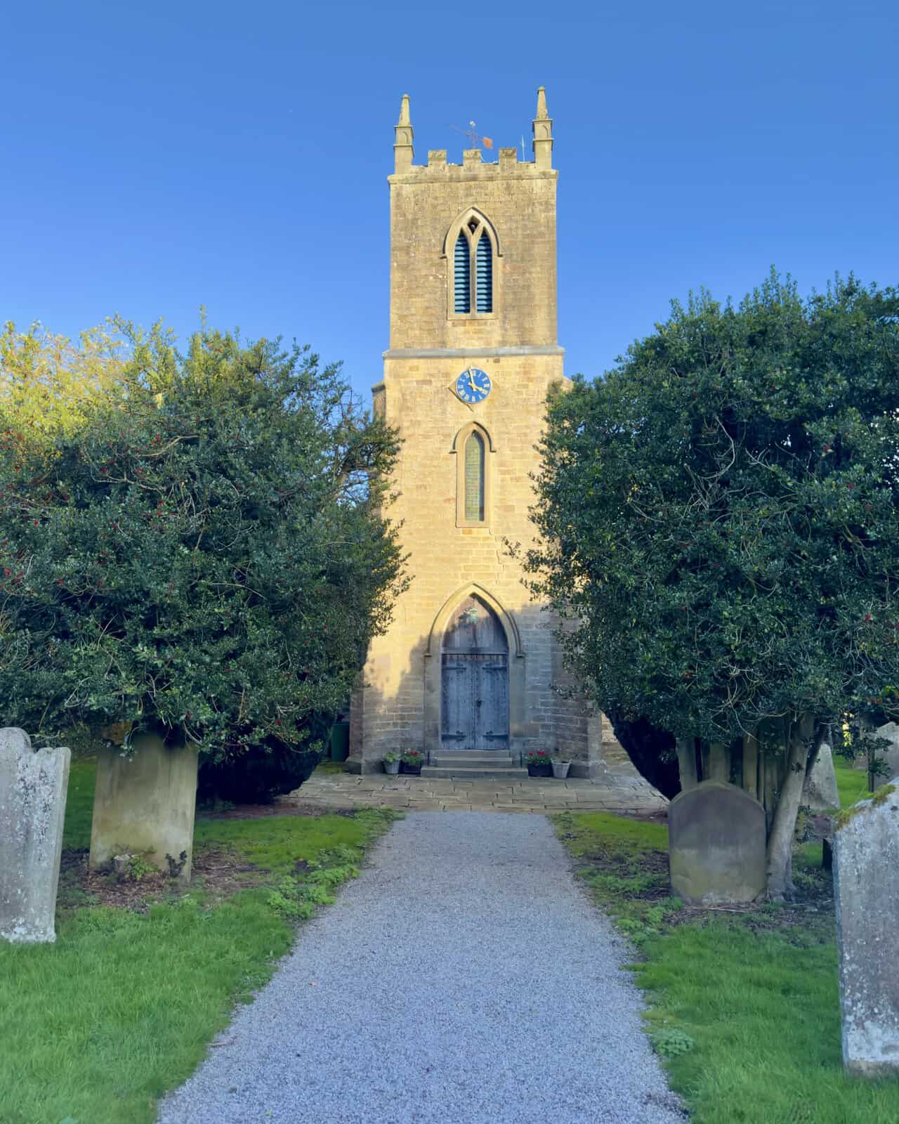 The tower of St Mary's Church, visited at the end of the Ramsgill walk, with corner pinnacles, a blue clock face and holly trees framing the path.