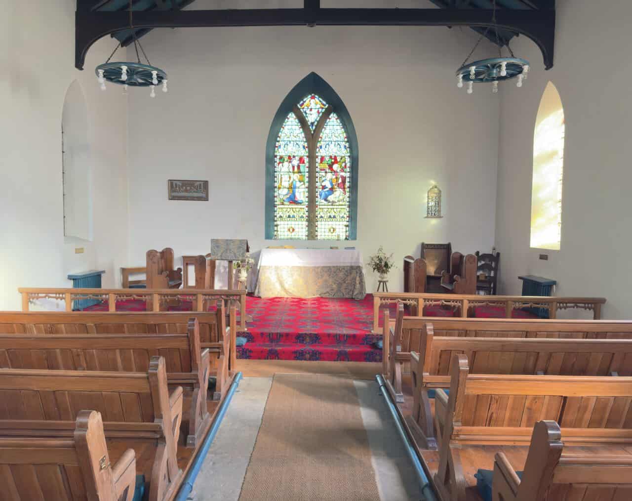 The simple interior of St Mary's Church, Ramsgill, with wooden pews, a red-carpeted sanctuary and a stained-glass window above the altar.